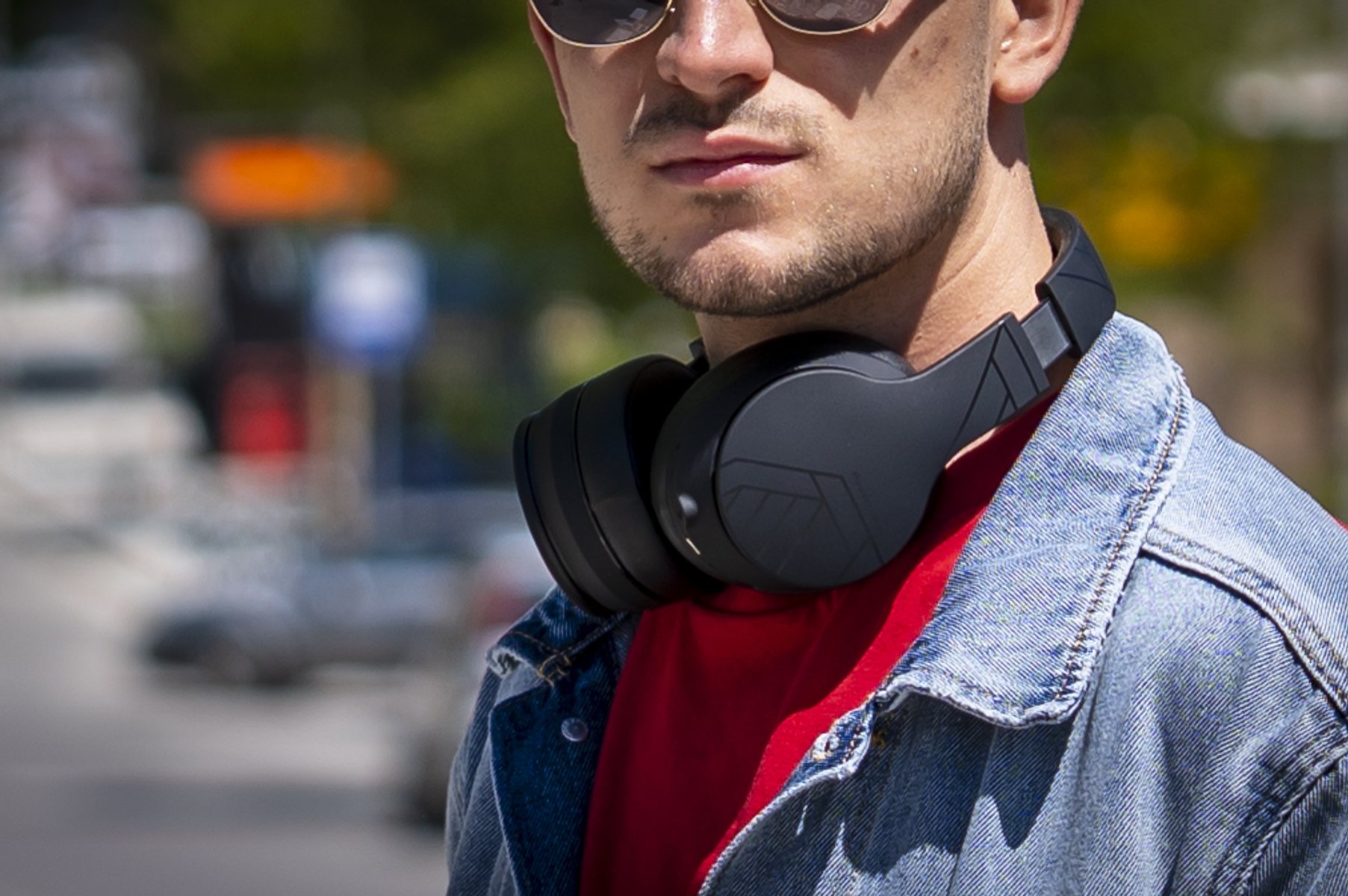 Un hombre con gafas de sol tiene auriculares alrededor del cuello. Lleva una camisa roja y una chaqueta vaquera azul. Fondo borroso.