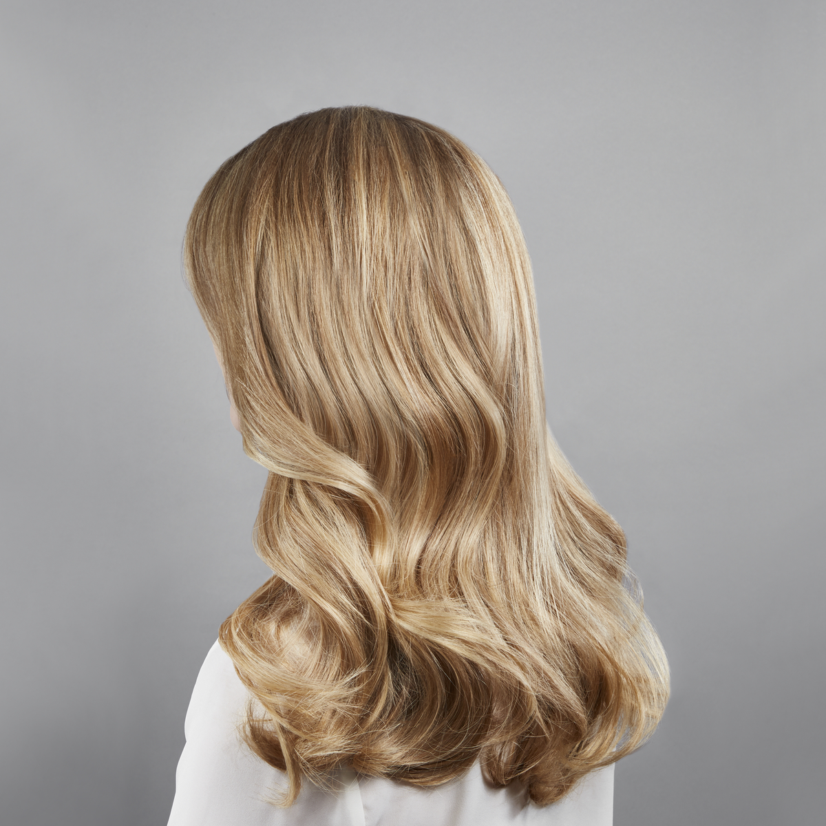 Osoba z długimi, falistymi blond włosami, widoczna od tyłu, na szarym tle. Ma na sobie białą bluzkę.