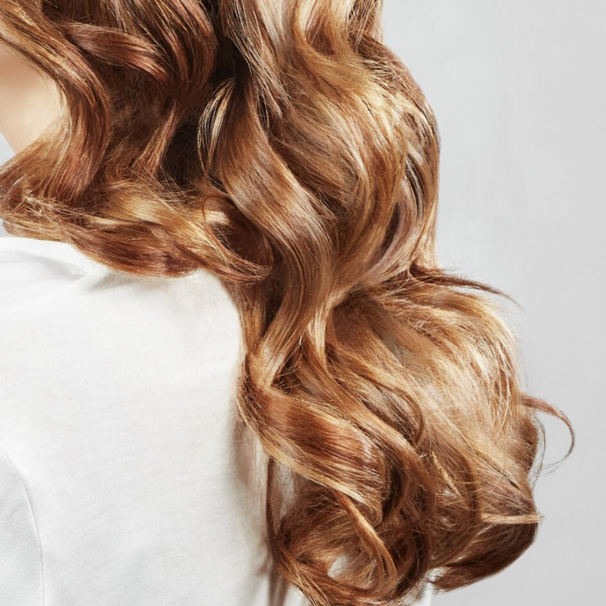 Zbliżenie falujących, kasztanowych włosów opadających na białą koszulę. Włosy są dobrze oświetlone, ukazując różne odcienie brązu i złota.