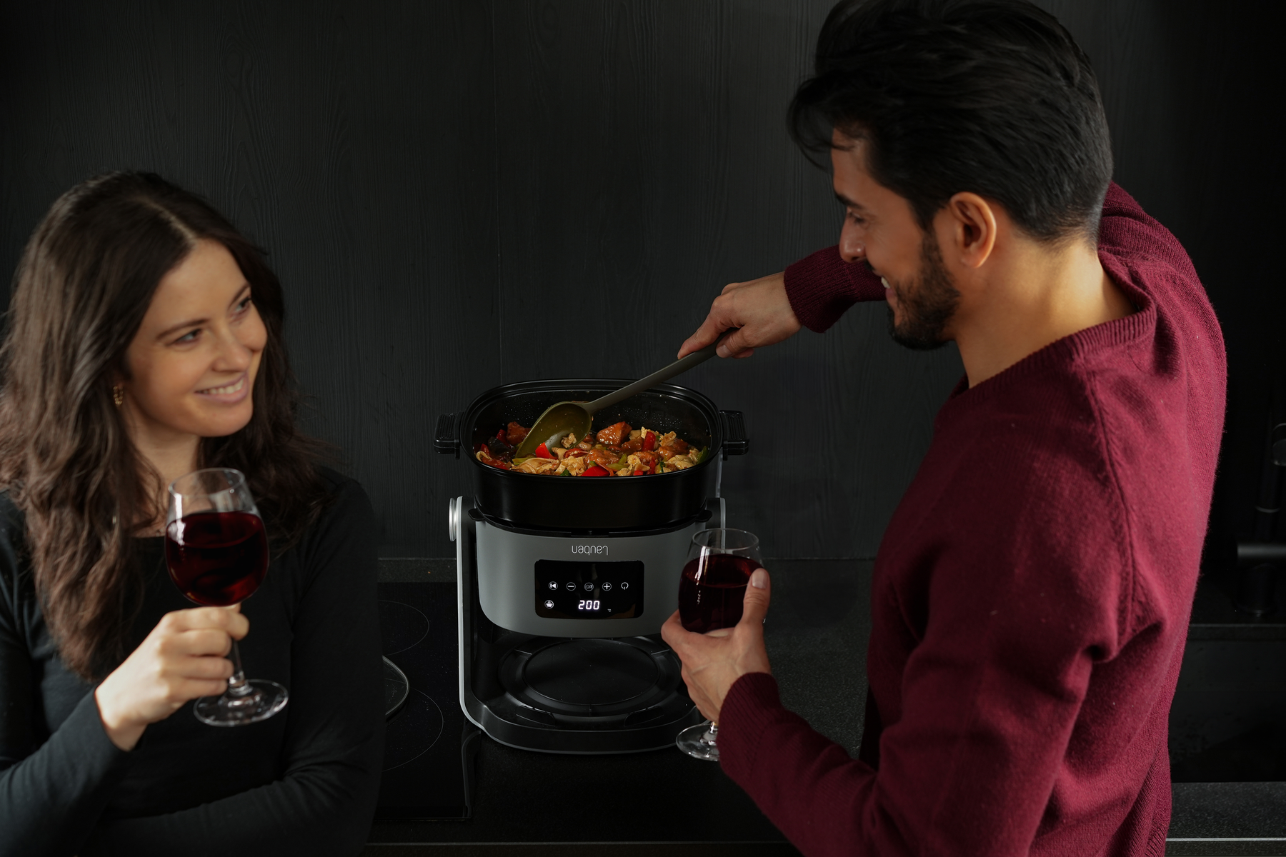 Mężczyzna miesza jedzenie w garnku, a kobieta trzyma kieliszek wina. Oboje się uśmiechają. Garnek stoi na blacie.