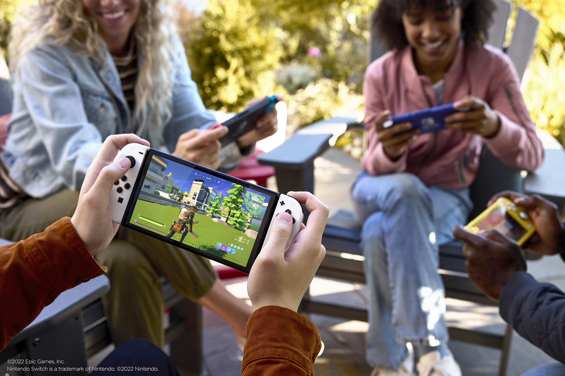 Persone che giocano ai videogiochi all'aperto. Mani che tengono un Switch. Altri giocano su dispositivi. Giorno di sole, alberi verdi sullo sfondo.