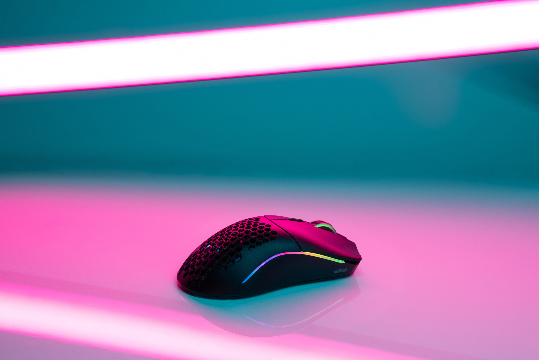 Eine schwarze Gaming-Maus mit RGB-Beleuchtung liegt auf einer reflektierenden Oberfläche. Der Hintergrund ist rosa und türkis mit hellem Deckenlicht.