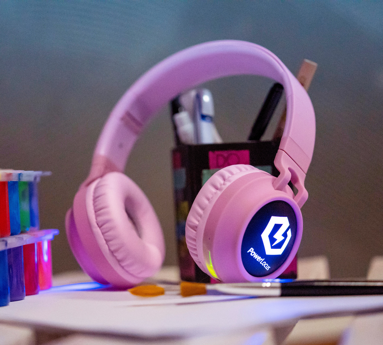 Cuffie rosa con il logo PowerLocus su un tavolo. Sono presenti anche tubetti di vernice, penne e un pennello.