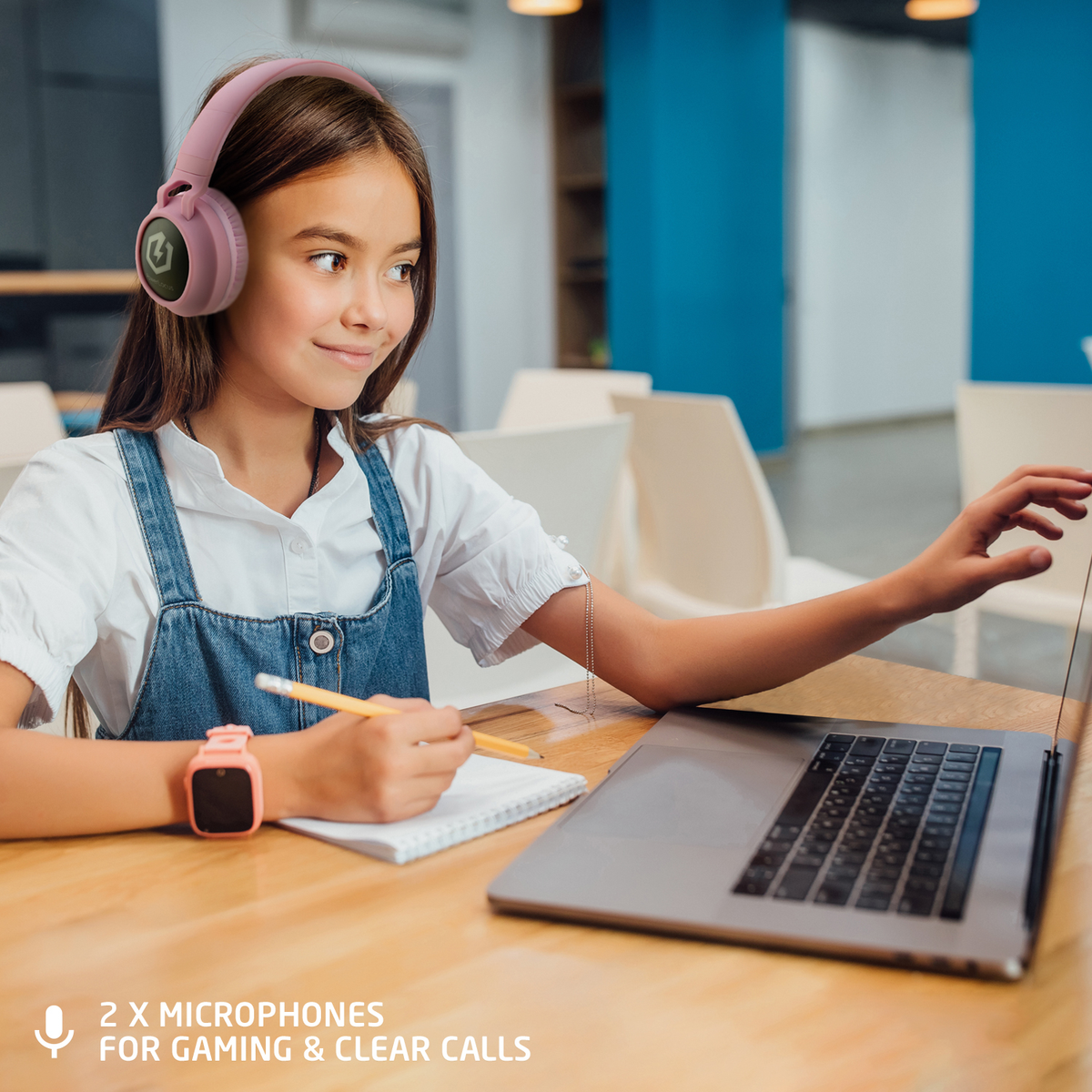Una ragazza con cuffie rosa, tuta in denim e smartwatch sorride mentre usa un laptop su un tavolo di legno.