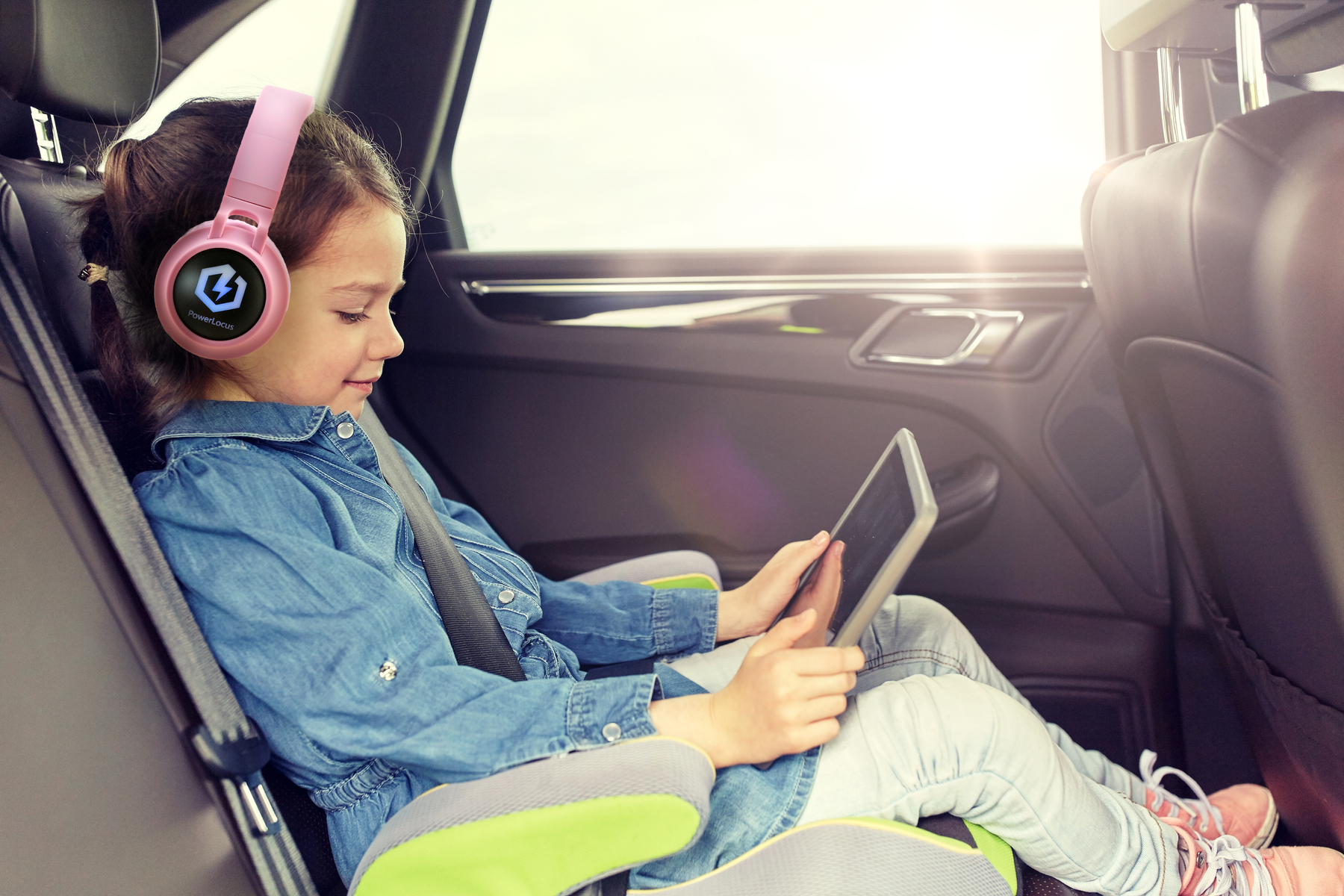 Ragazza in auto con cuffie rosa, usa un tablet. La luce del sole entra dalla finestra. Indossa una giacca di jeans e jeans.