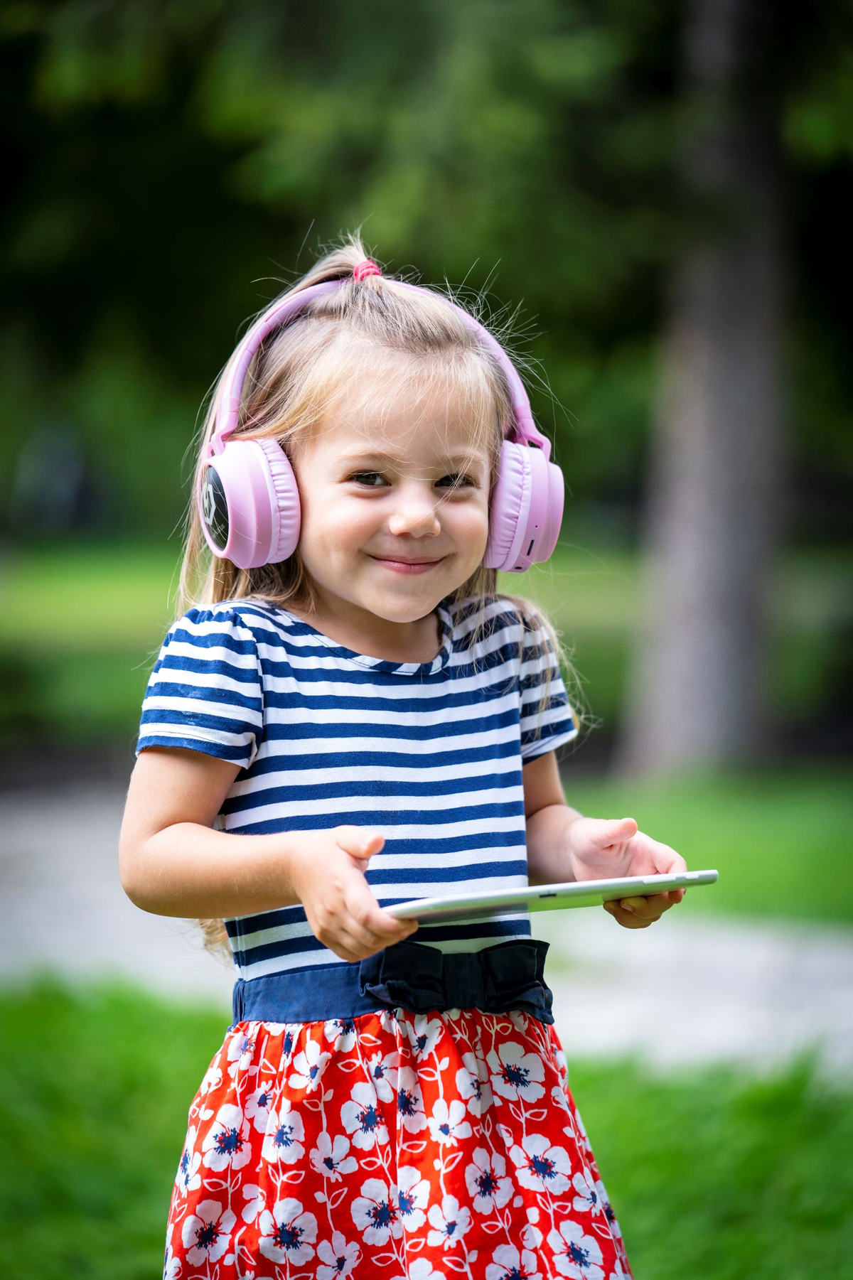 Una bambina sorridente con le cuffie tiene in mano un tablet. Indossa una maglietta a righe e una gonna a fiori. Erba verde e alberi sullo sfondo.