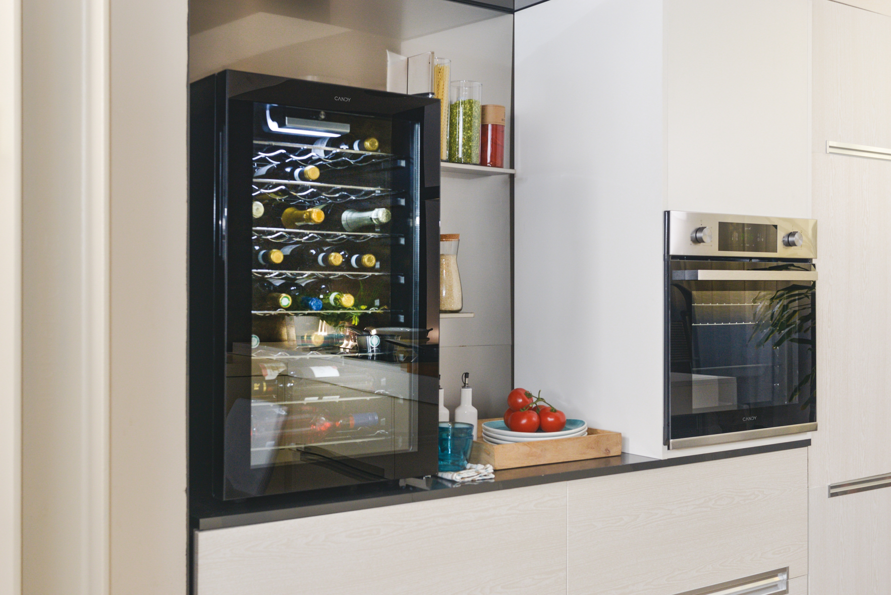 Una cucina con un frigo per vini pieno di bottiglie, un forno, mensole con barattoli e pomodori su un bancone.