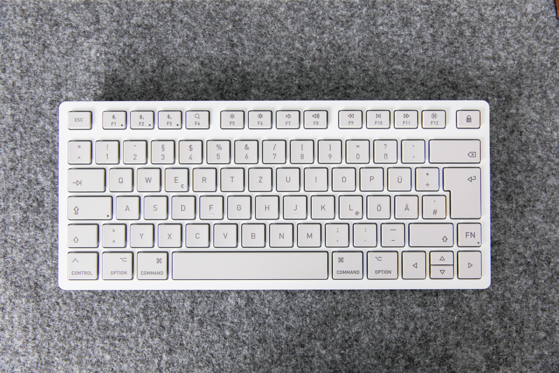 Teclado blanco sobre una superficie gris. Teclas con letras, números y símbolos visibles. El teclado está centrado.