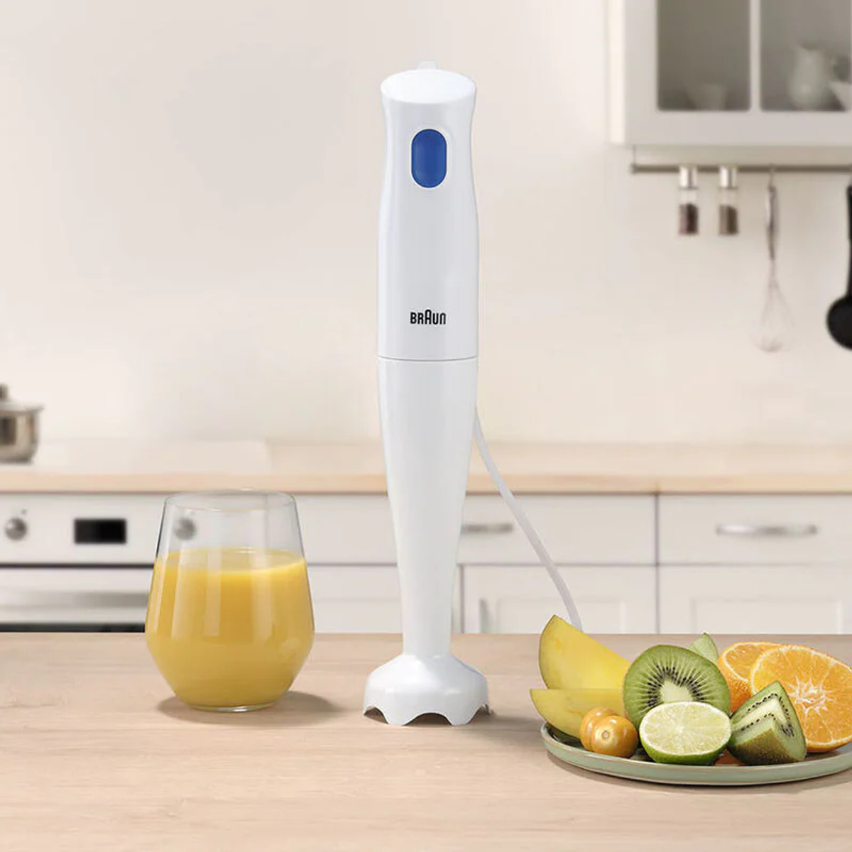 Ein weißer Braun-Stabmixer steht neben einem Glas Saft und einem Teller Obst auf einem Holztisch in einer Küche.