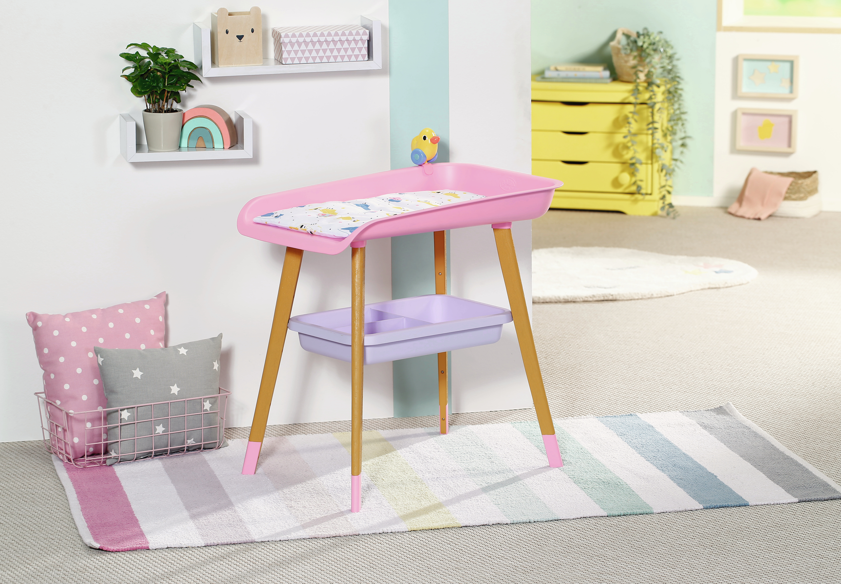 Cambiador rosa con patas de madera. Cojines rosas y grises en una cesta. Alfombra a rayas, estanterías con decoración al fondo.