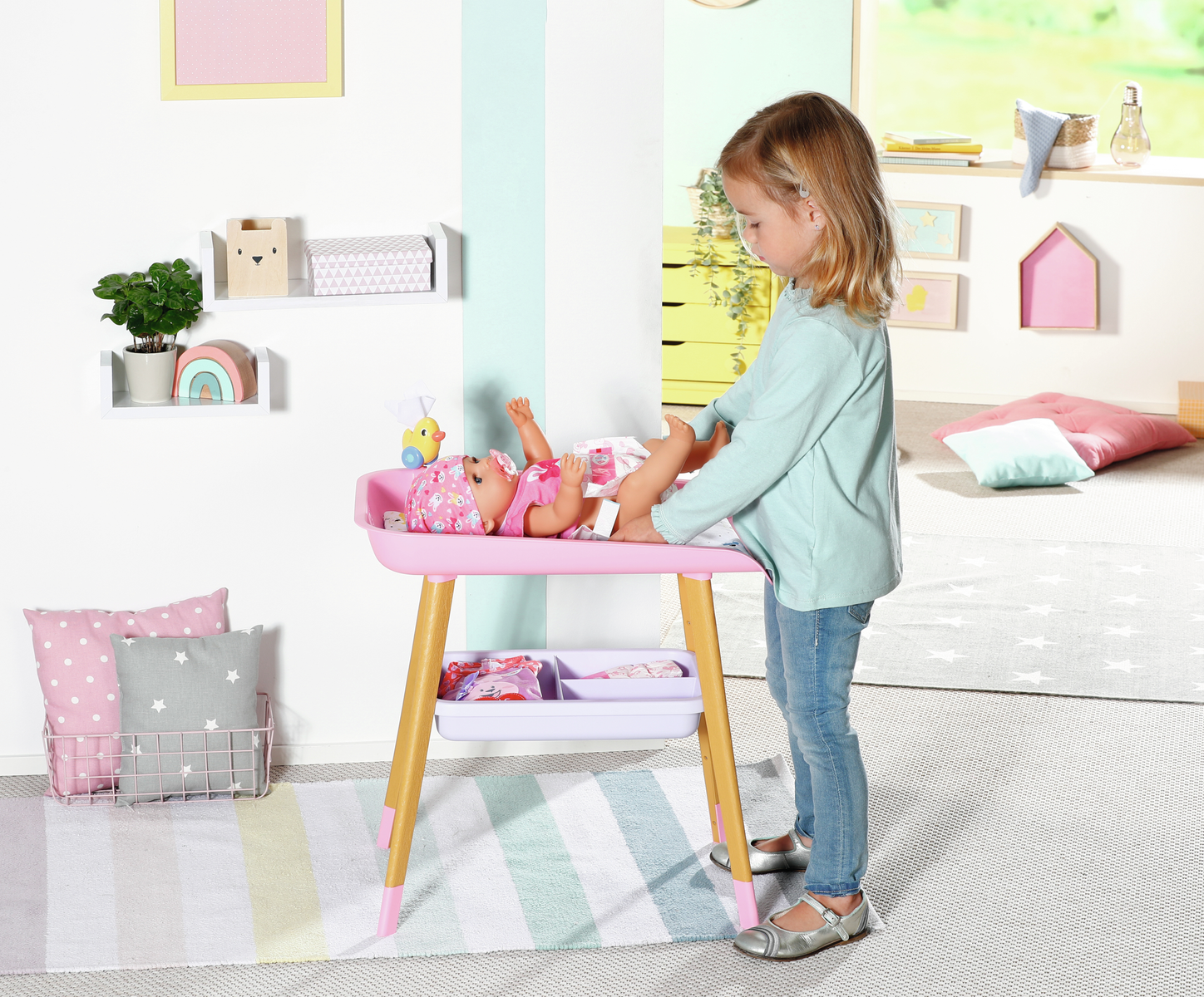 Una niña está de pie junto a un cambiador rosa con una muñeca. La habitación tiene estanterías, una alfombra y una ventana. La niña está cambiando el pañal de la muñeca.