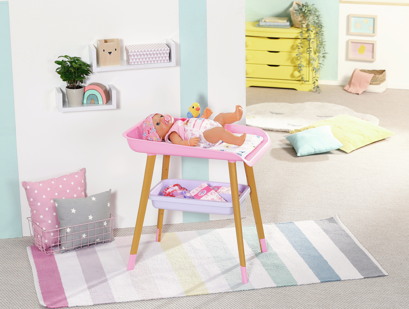 Una muñeca bebé está en una mesa cambiador rosa. Hay estantes con decoración, almohadas y una alfombra en la habitación. Una cómoda amarilla está detrás.