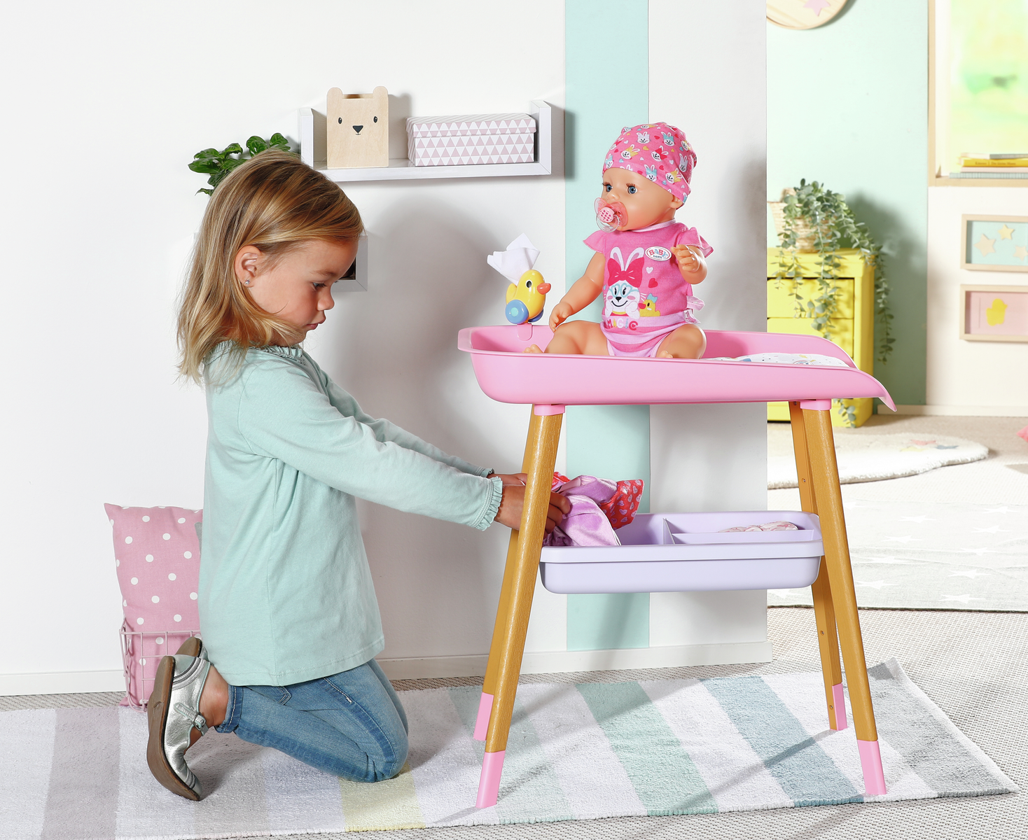 Una niña se arrodilla junto a un cambiador de muñecas rosa con una muñeca con un conjunto rosa. Una estantería y un juguete están sobre la mesa.