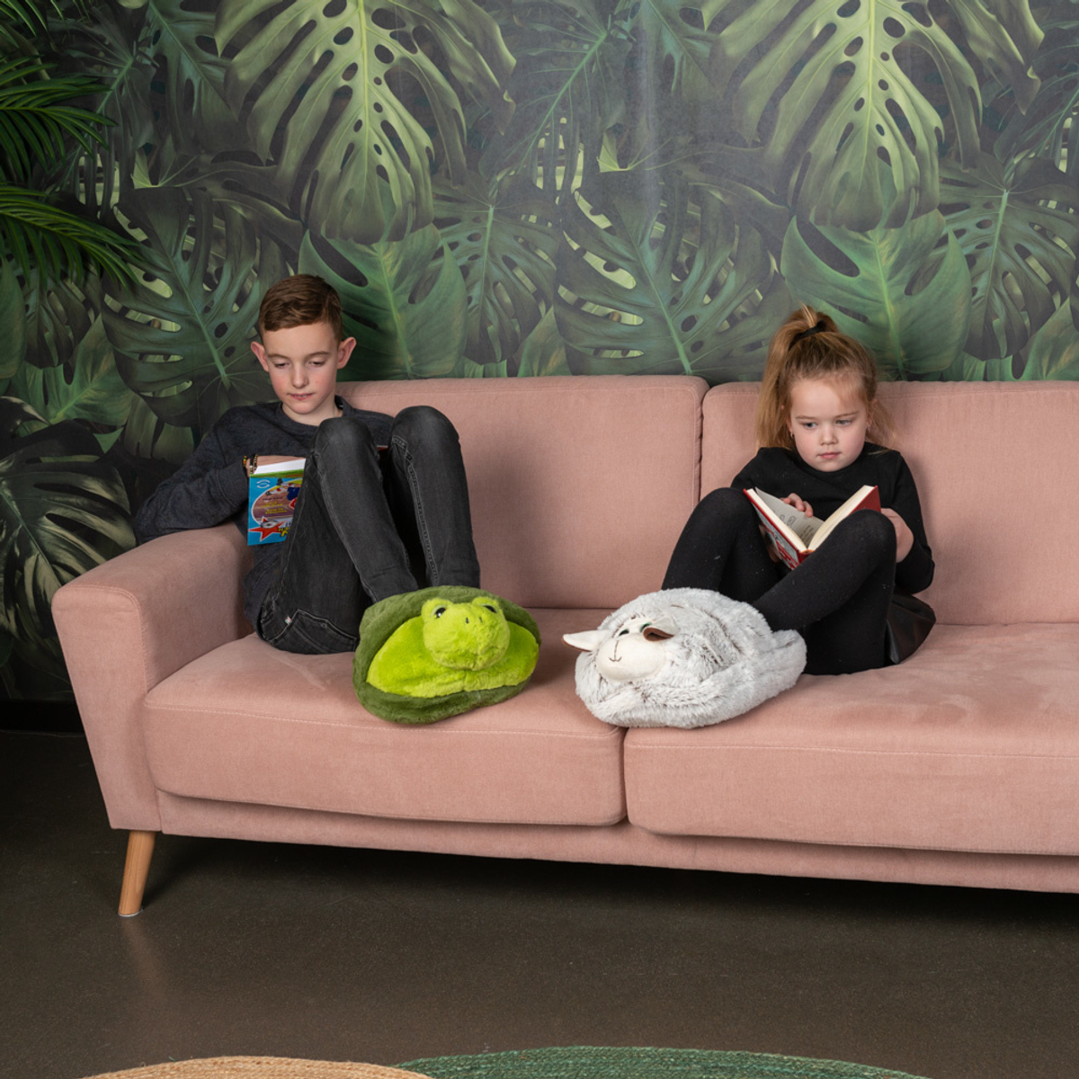 Zwei Kinder sitzen auf einem rosa Sofa und lesen Bücher. Einer trägt Schildkrötenslipper, der andere Schafslippers, vor einer Wand mit Blattmuster.