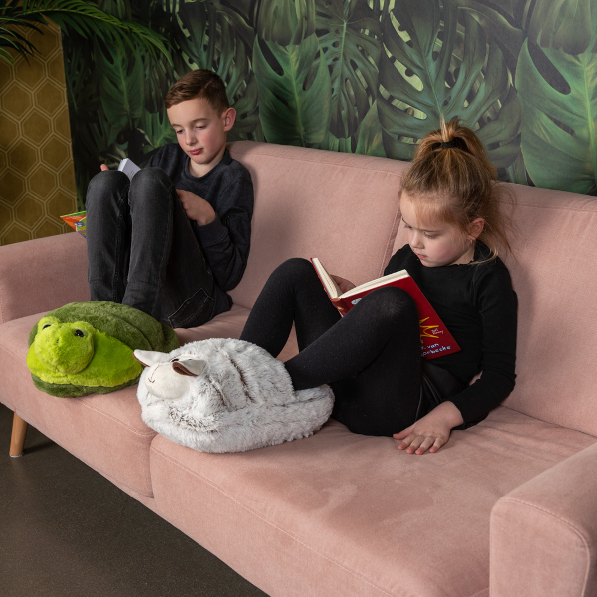 Zwei Kinder lesen Bücher auf einem rosa Sofa, ein grüner Frosch und ein weißes Schaf-Plüschtier sind vor ihnen. Tropische Tapete im Hintergrund.