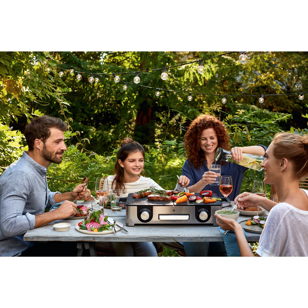Rodzina cieszy się grillem. Kobieta nalewa wodę z dzbanka. Grill z jedzeniem. Ludzie uśmiechają się i jedzą przy drewnianym stole.