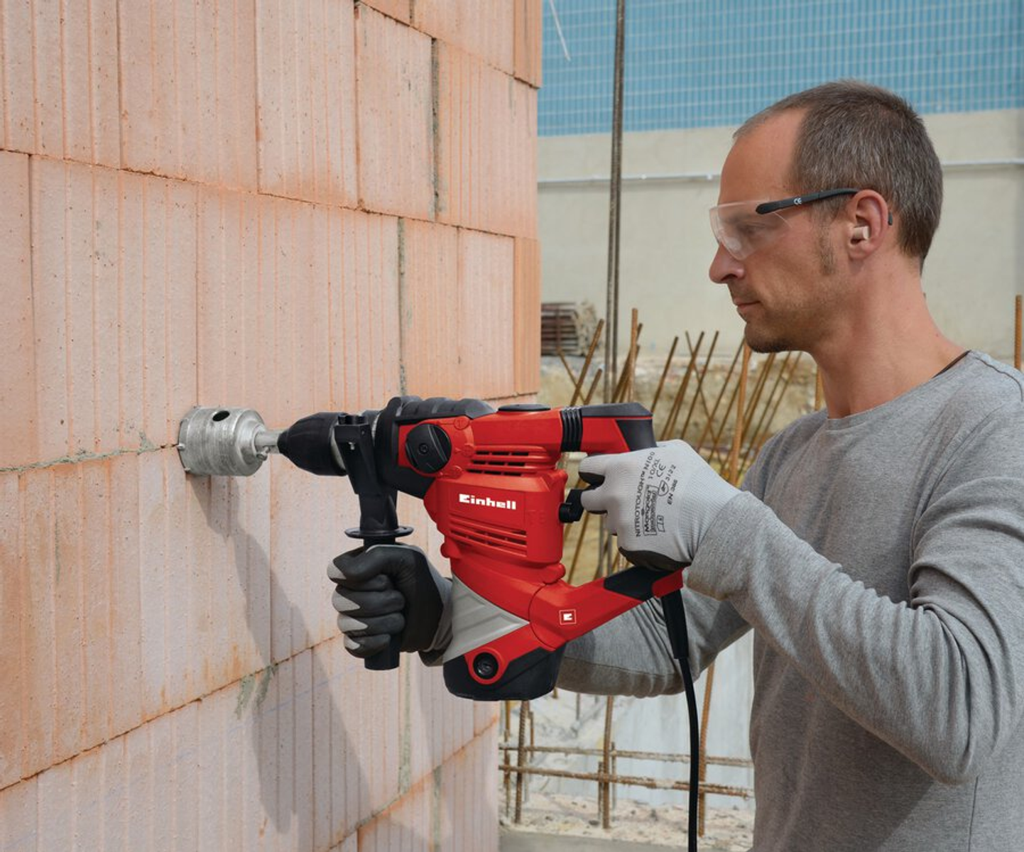 Un hombre taladra un agujero en una pared de ladrillos con un taladro rojo. Usa gafas y guantes de seguridad. Obra en el fondo.