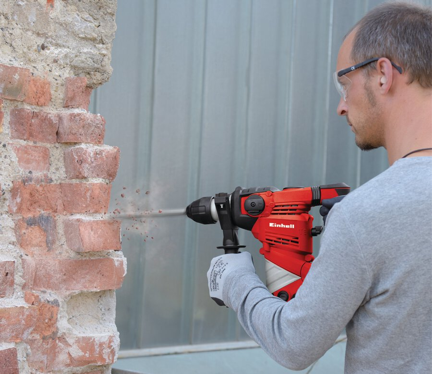Un hombre taladra una pared de ladrillo con un taladro rojo Einhell. Lleva gafas de seguridad y guantes. La pared se está desmoronando.