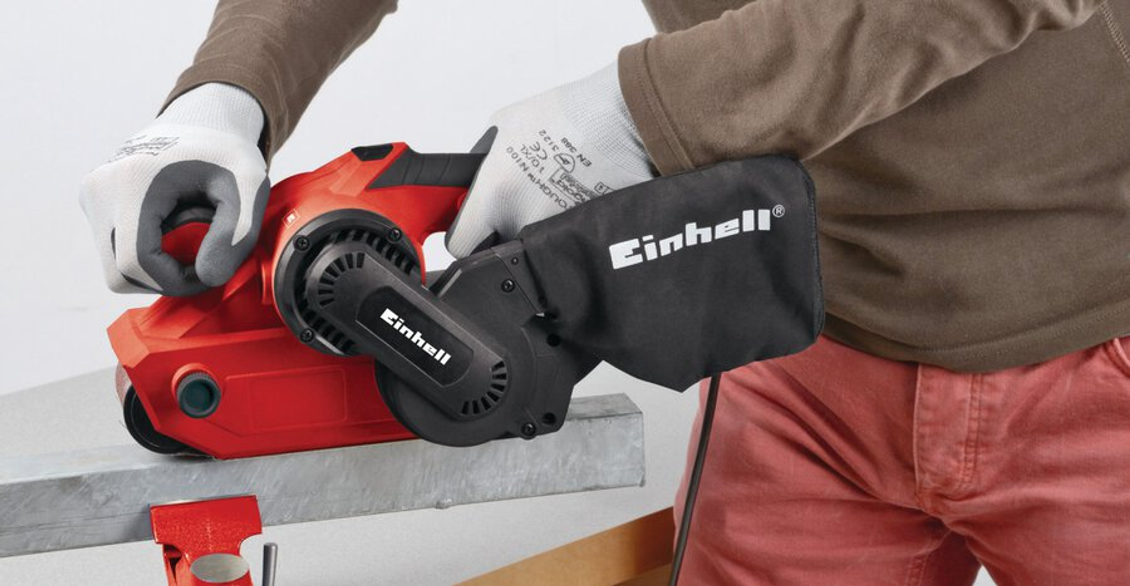 Une personne utilise une ponceuse Einhell rouge sur du métal. Elle porte des gants et une chemise marron, avec un sac à poussière noir attaché.
