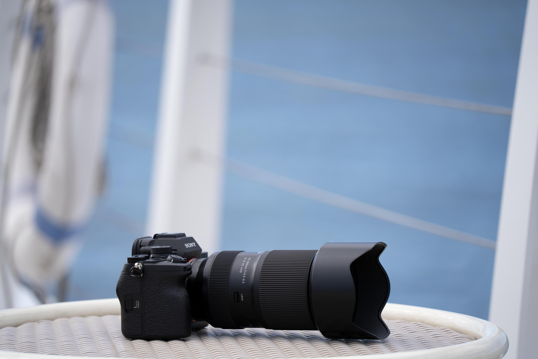 Czarny aparat z obiektywem leży na białym stole. Tło to rozmyty widok niebieskiego nieba i balustrady łodzi.