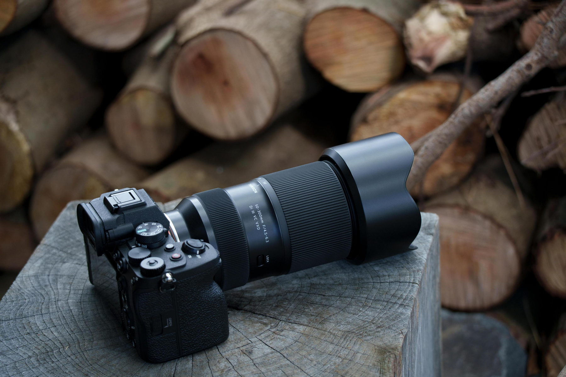 Czarny aparat z obiektywem na drewnianej powierzchni, kłody w tle. Widoczne detale aparatu: TAMRON 50-300mm F/4.5-6.3.