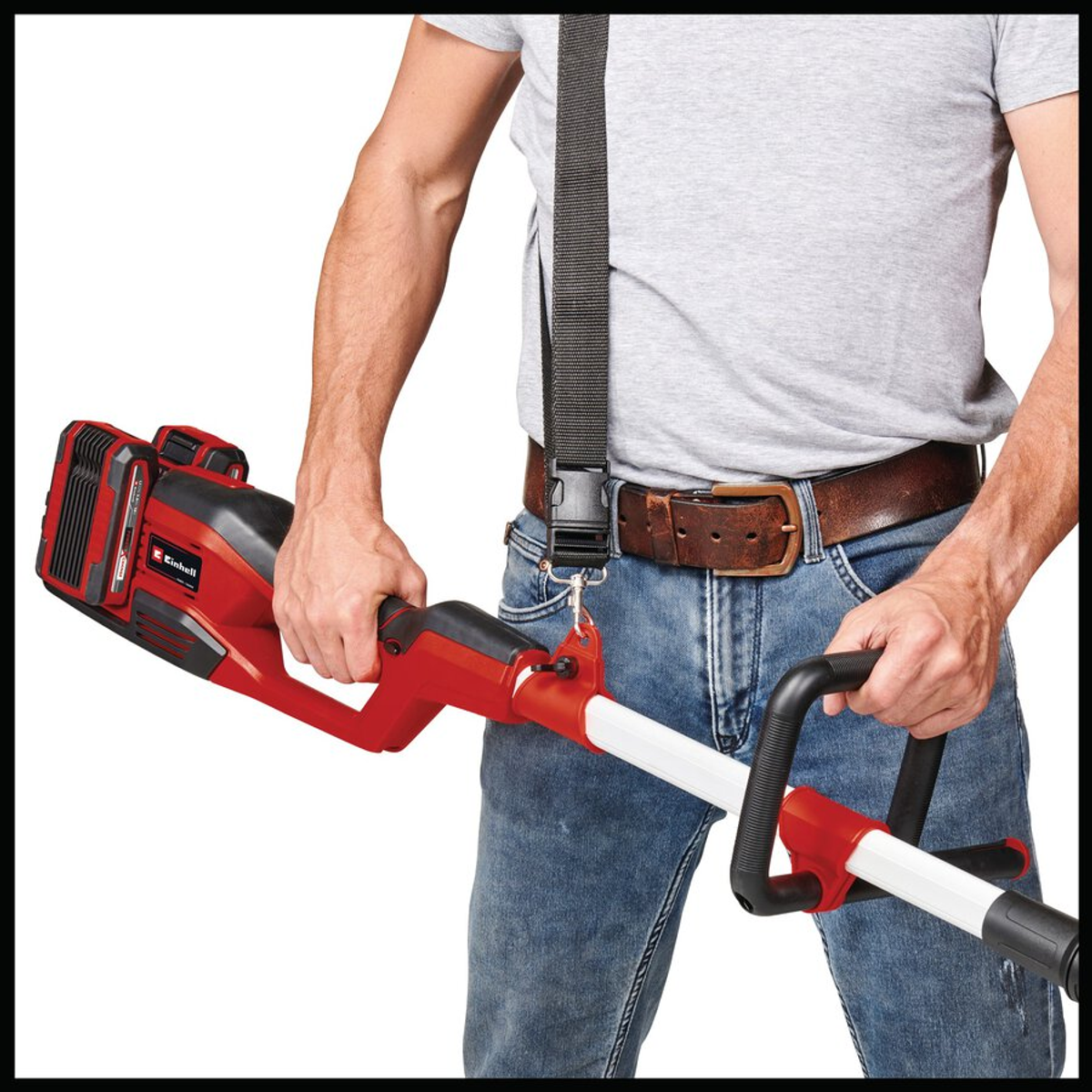 Hombre sosteniendo un cortador Einhell rojo y negro. Lleva vaqueros, una camisa gris y una correa negra. Se ve un cinturón marrón.