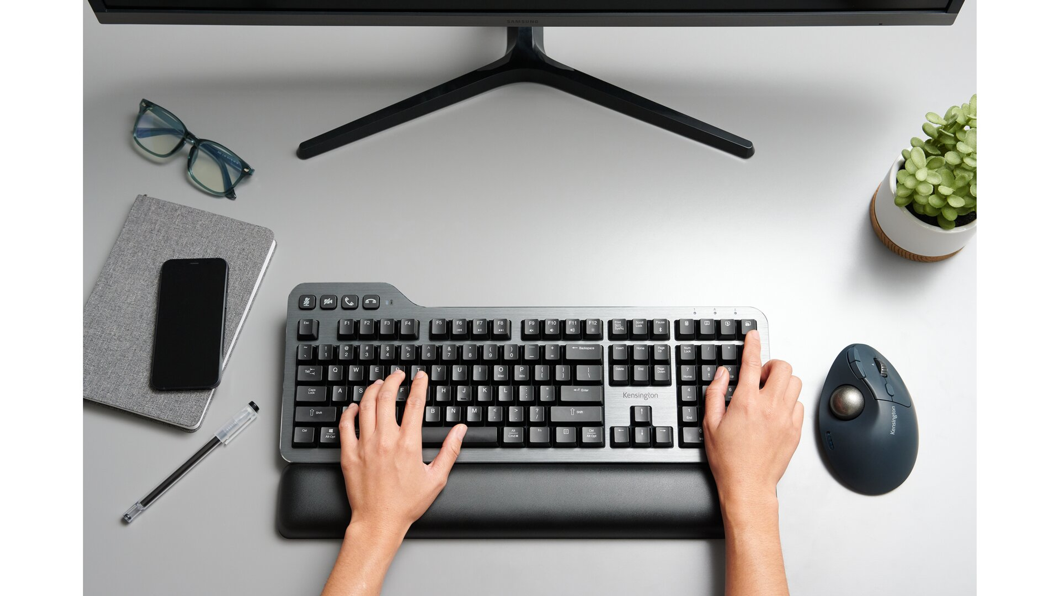 Overhead view of a desk with keyboard, mouse, monitor, glasses, notebook, and pen. Hands are on the keyboard.