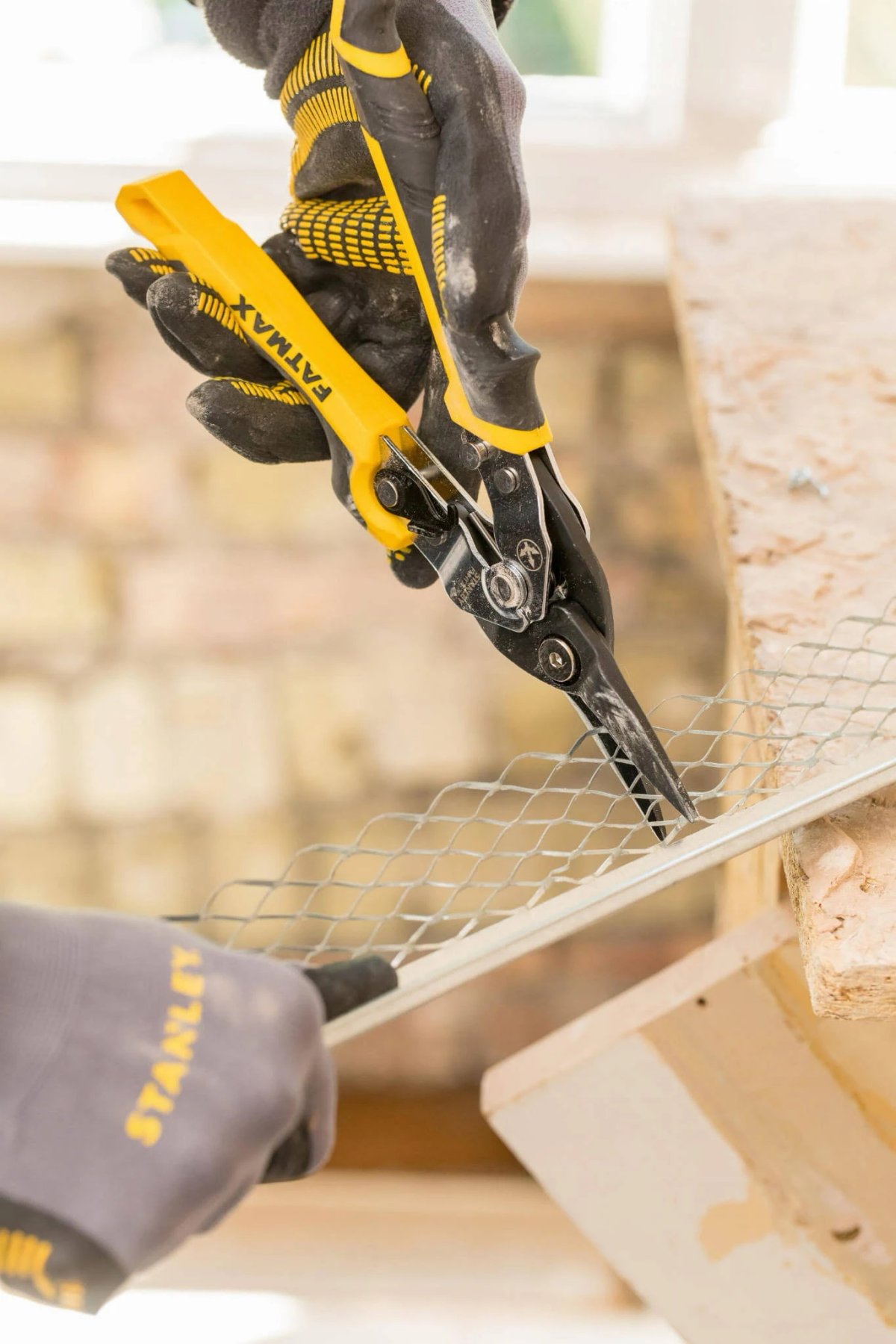 Eine Person mit Handschuhen schneidet mit einer gelb-schwarzen Schere Metallgitter. Das Gitter befindet sich auf einer Holzkonstruktion.