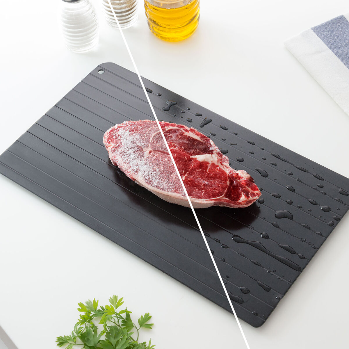 Rohes Steak auf schwarzem Tablett, halb gefroren. Öl, Salz, Pfeffer und Petersilie sind in der Nähe. Weißer Hintergrund. Wassertropfen um das Tablett herum.