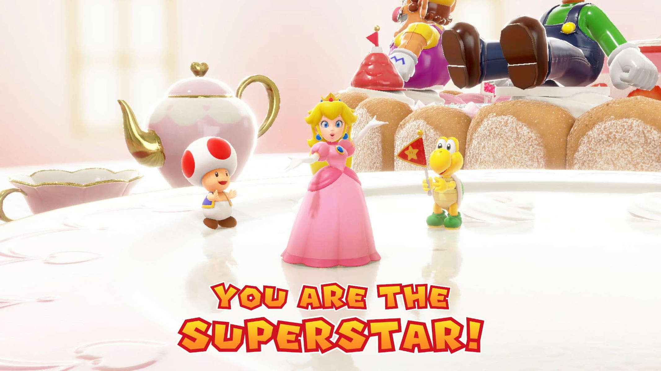 Peach, Toad e Koopa Troopa sono su una torta. Wario e Luigi sono sulla torta sullo sfondo. Testo: SEI LA SUPERSTAR!