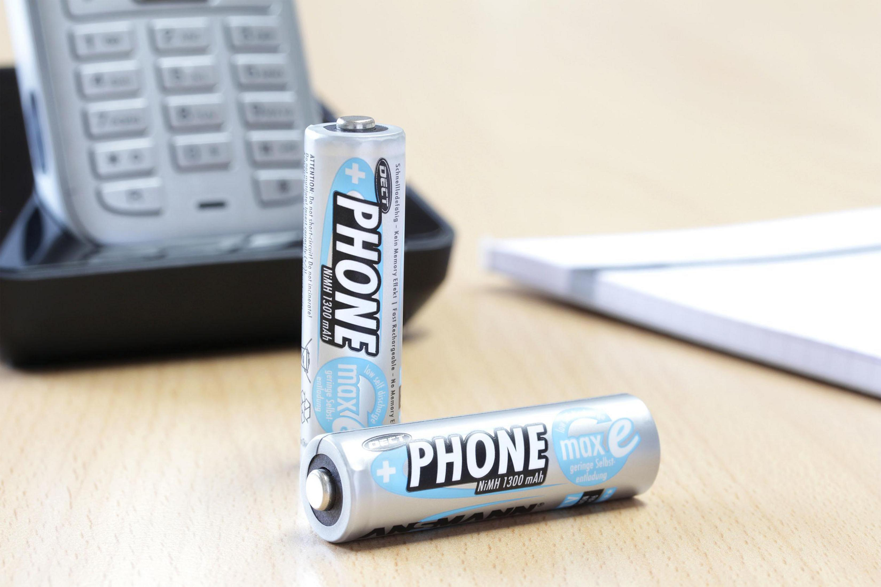 Dos pilas plateadas PHONE sobre una superficie de madera, base de teléfono y bloc de notas en el fondo.