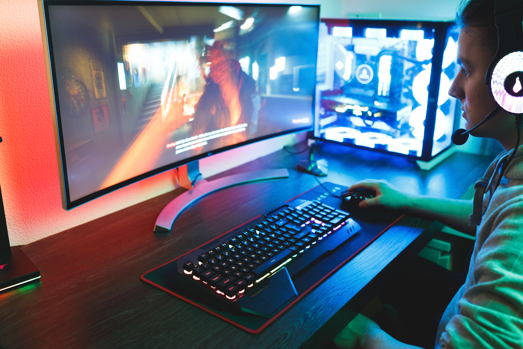 Osoba z zestawem słuchawkowym gra na komputerze. Ekran wyświetla scenę z gry. Klawiatura i mysz są na biurku.