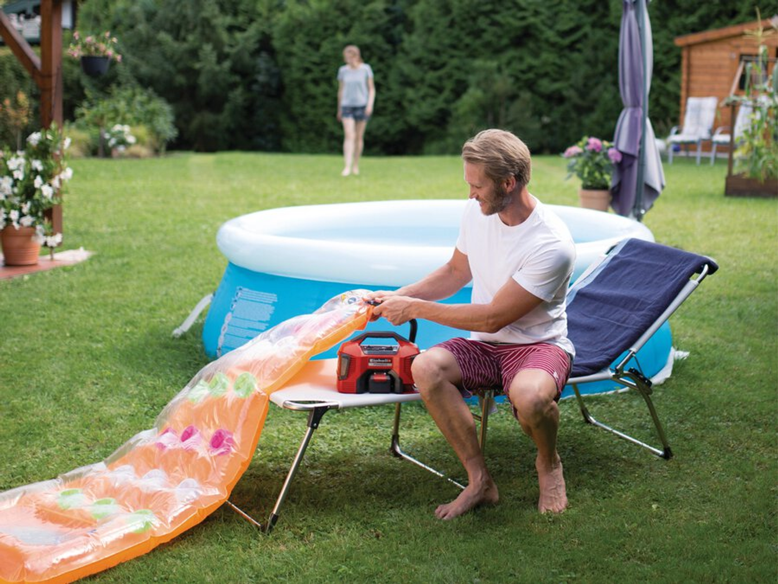 Ein Mann pumpt ein Pool-Schwimmteil auf. Im Hintergrund sind ein Pool und eine Frau. Grünes Gras umgibt die Szene.
