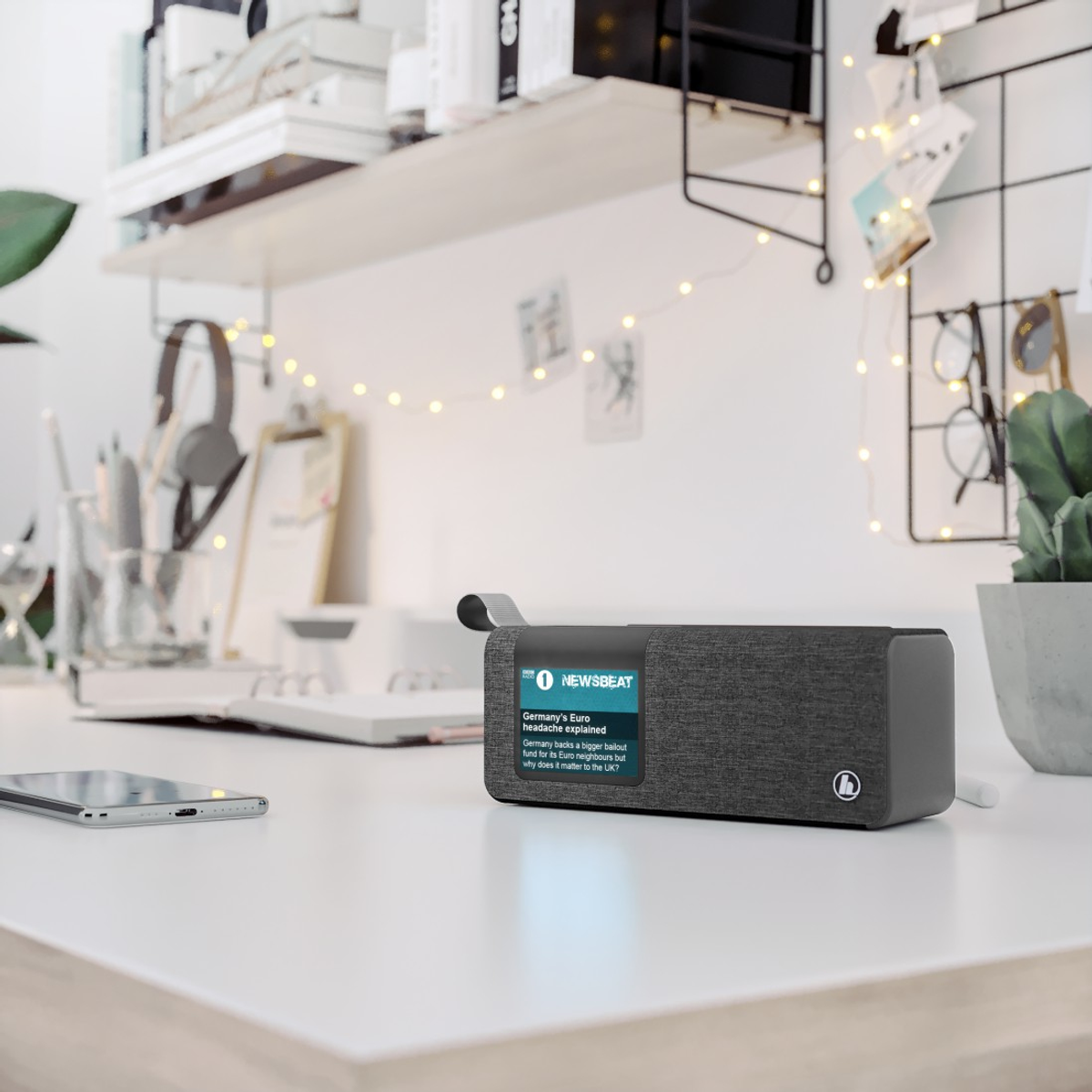 Un escritorio con un altavoz inteligente mostrando noticias, un teléfono y útiles de oficina. Una estantería con libros y luces decorativas en el fondo.