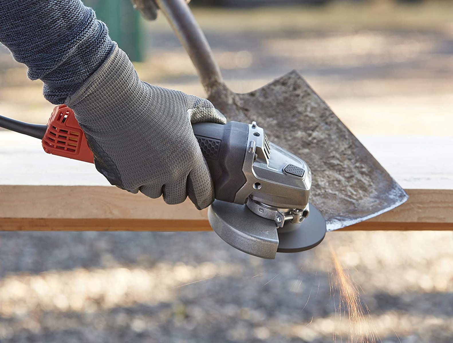 Een persoon slijpt hout met een haakse slijper. Vonken vliegen. Een schop staat op de achtergrond. De persoon draagt handschoenen.