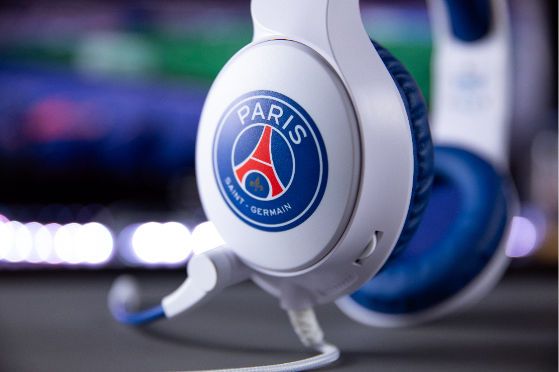 Zbliżenie białych i niebieskich słuchawek z logo Paris Saint-Germain. Tło jest rozmyte, z światłami i monitorem.