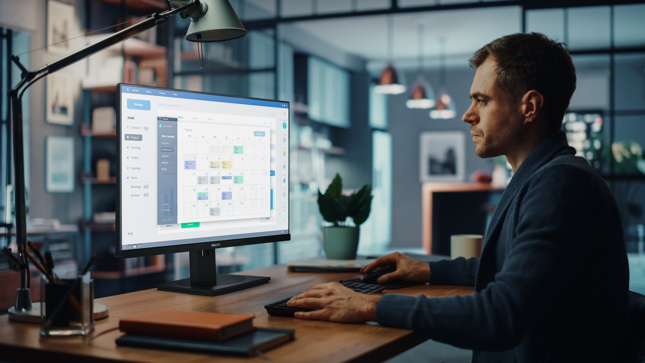 Mężczyzna siedzi przy biurku i patrzy na ekran komputera. Na ekranie wyświetlany jest kalendarz. Jest lampa, notatnik i roślina.