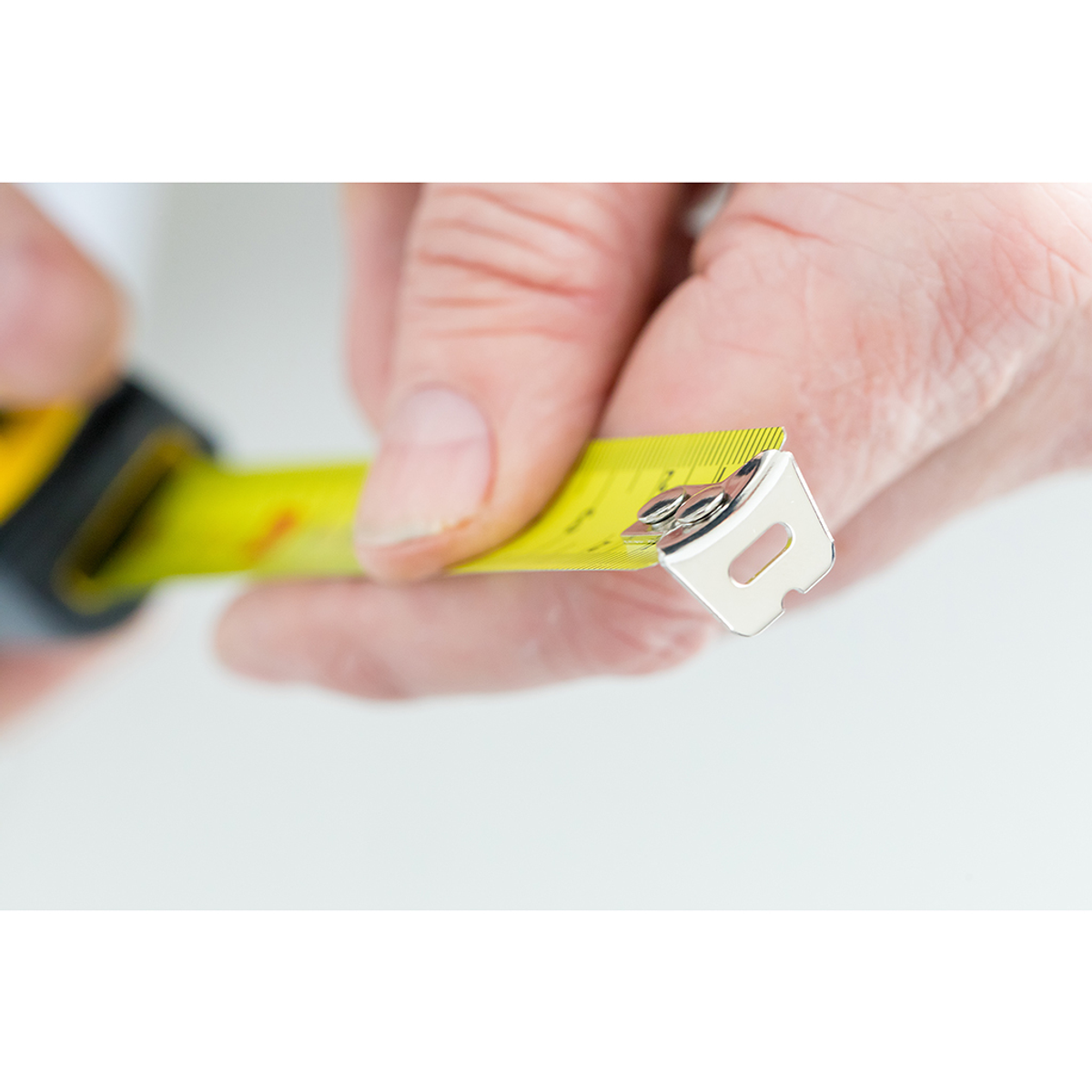 Ręka trzyma żółtą miarkę. Taśma jest wyciągnięta, koniec jest ostry. Tło jest białe.