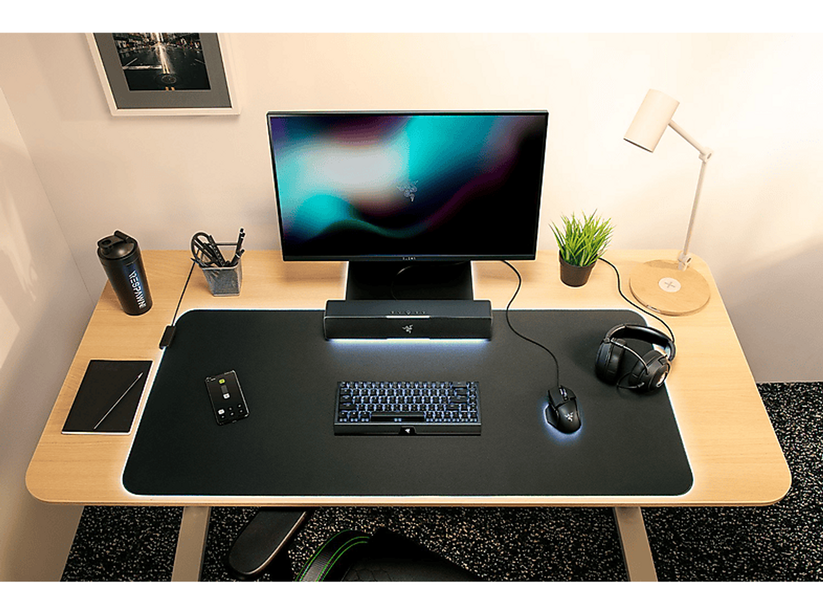 Biurko do gier z monitorem, klawiaturą, myszką, słuchawkami i rośliną. Widoczna jest lampa i butelka z wodą.