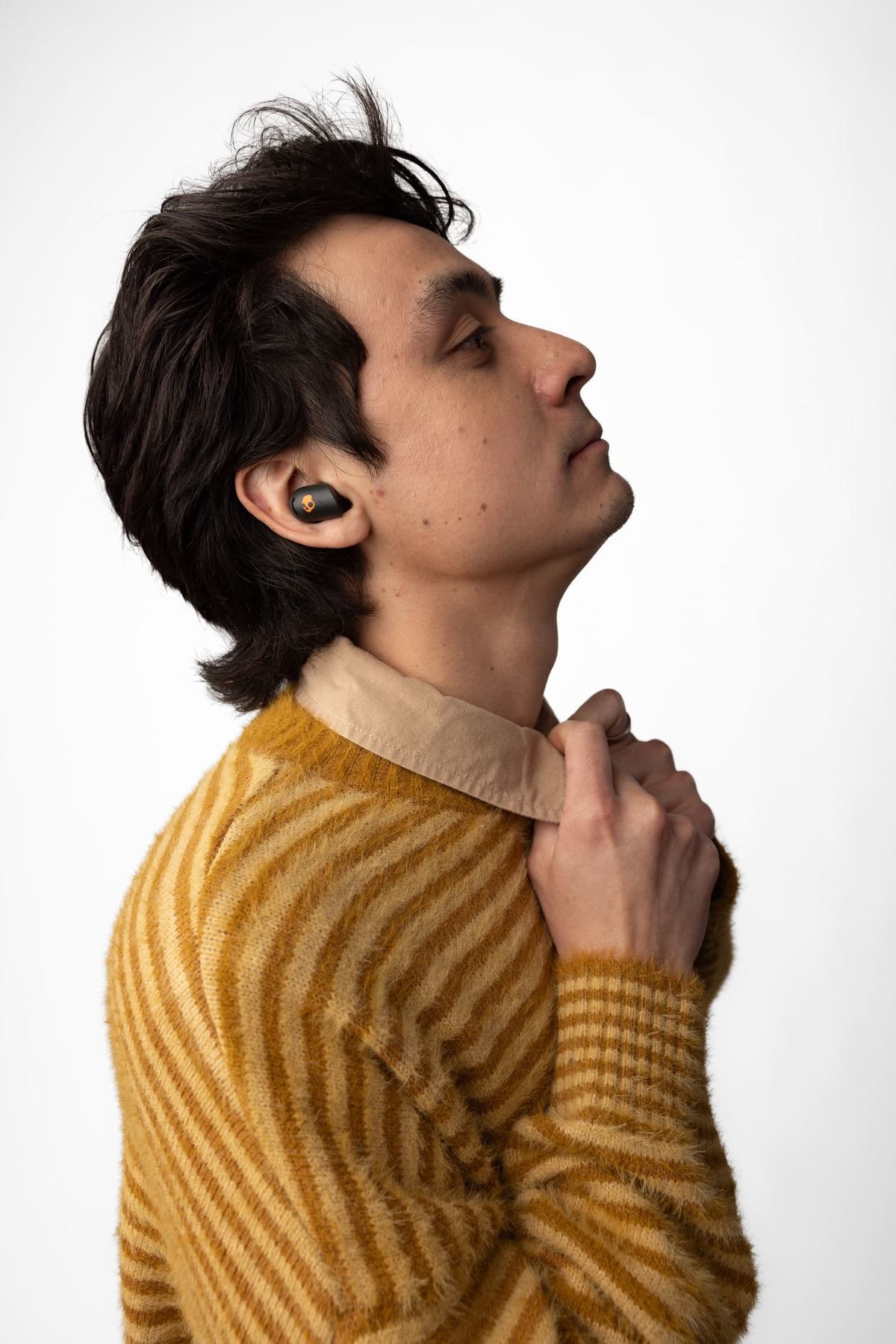 Mężczyzna z ciemnymi włosami w złotym swetrze trzymający kołnierz. Ma słuchawkę w uchu i patrzy w górę.