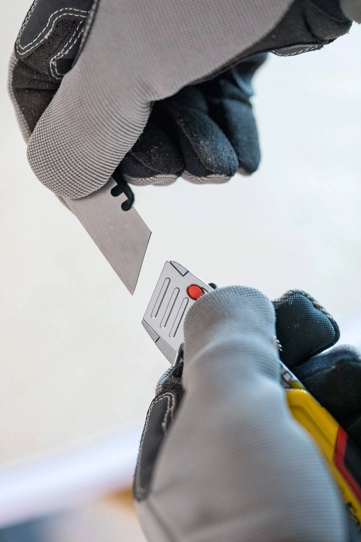 Hände mit Handschuhen halten ein Gebrauchsmesser, Klinge vom Griff getrennt. Roter Knopf am Griff. Grau- und Schwarztöne dominieren.