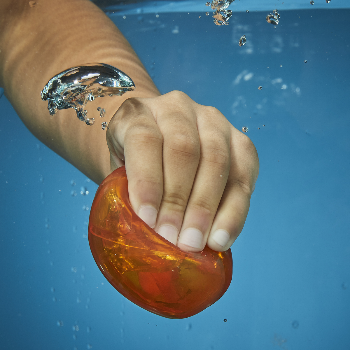 Dłoń trzyma pomarańczowy, przezroczysty przedmiot pod wodą. Pęcherzyki powietrza unoszą się wokół dłoni i przedmiotu. Tło to niebieska woda.