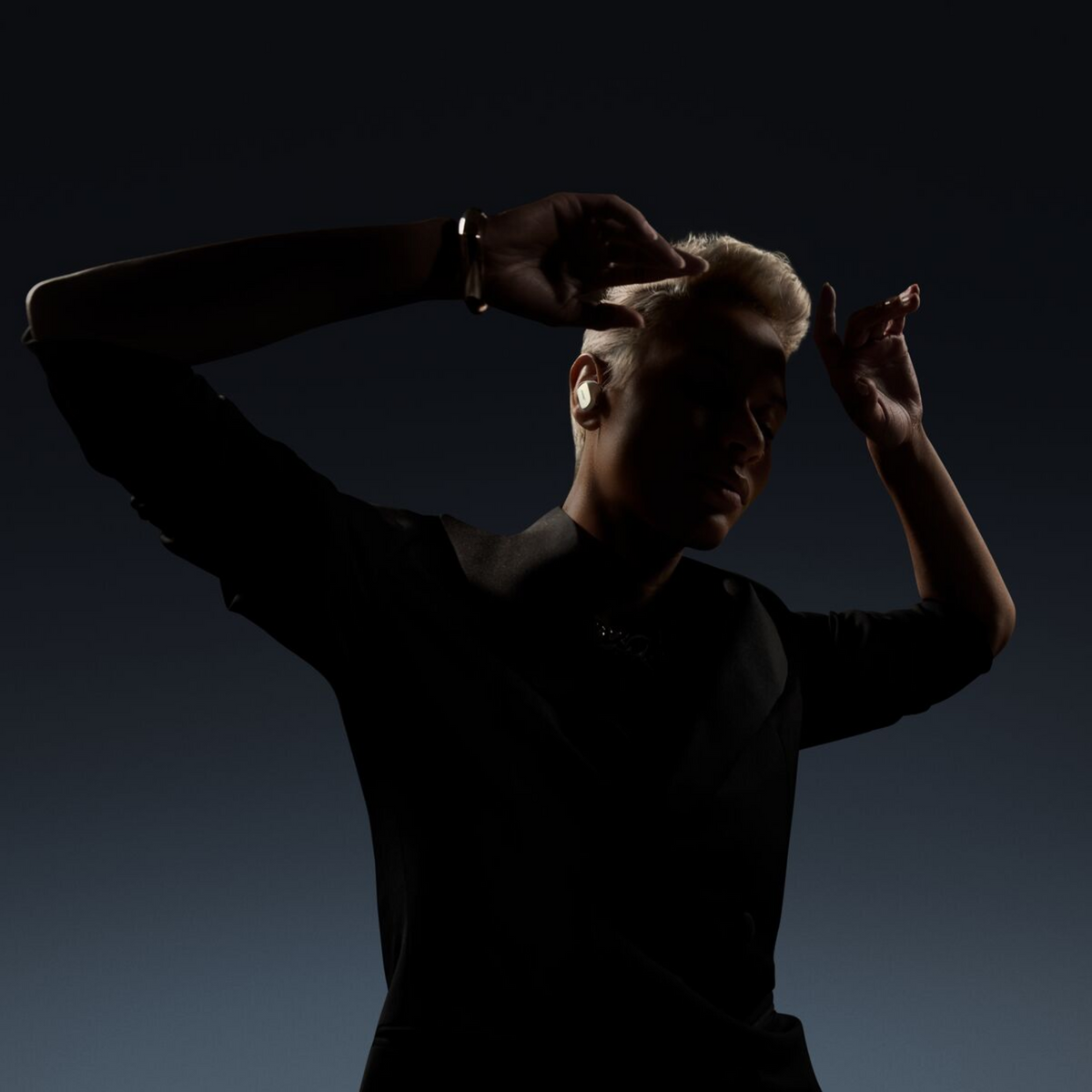 Osoba w sylwetce nosi słuchawki i podnosi ramiona, dłonie blisko głowy. Widoczne jasne włosy i bransoletka. Ciemne tło.