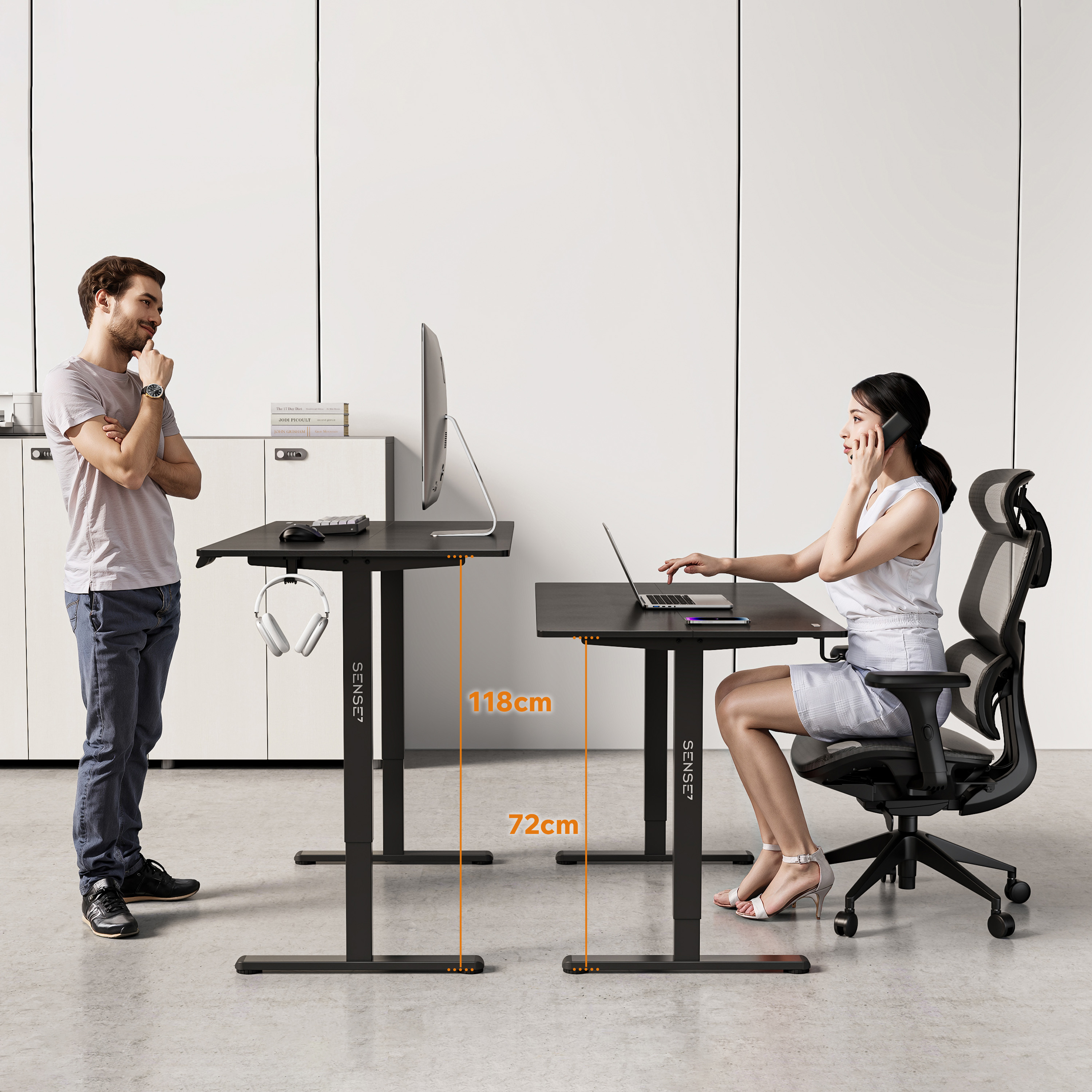 Ein Mann und eine Frau an Schreibtischen. Die Frau sitzt und telefoniert an einem Schreibtisch mit Laptop.