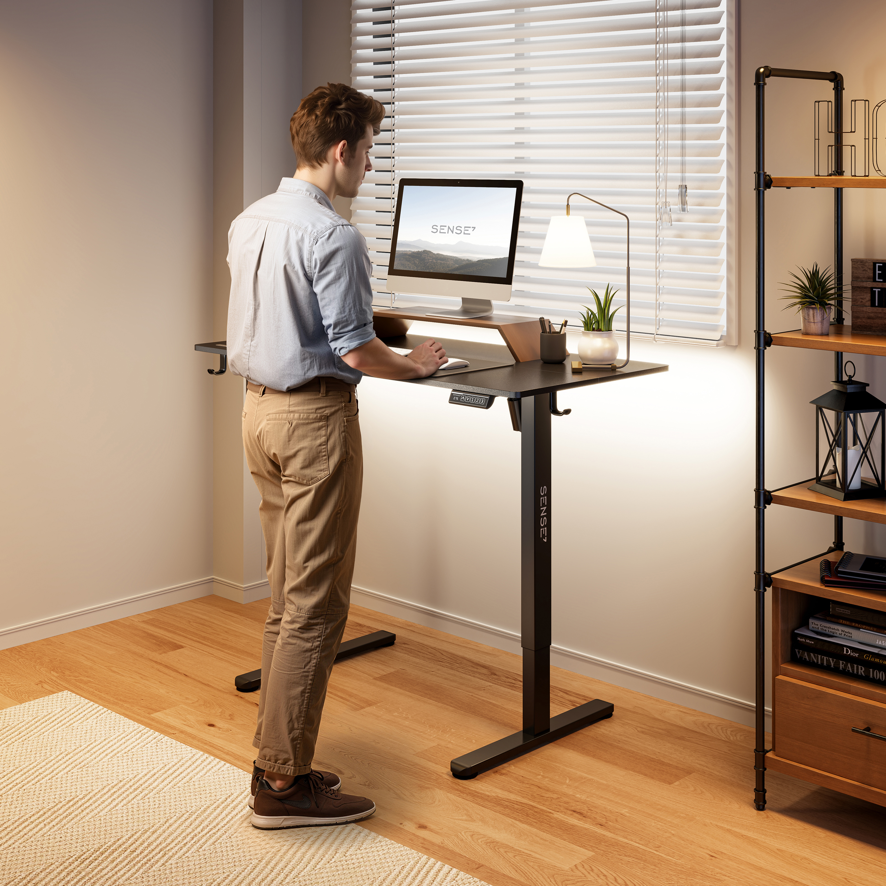 Een persoon staat aan een zwart verstelbaar bureau met een computer. Een lamp en planken zijn rechts. Het raam heeft jaloezieën.