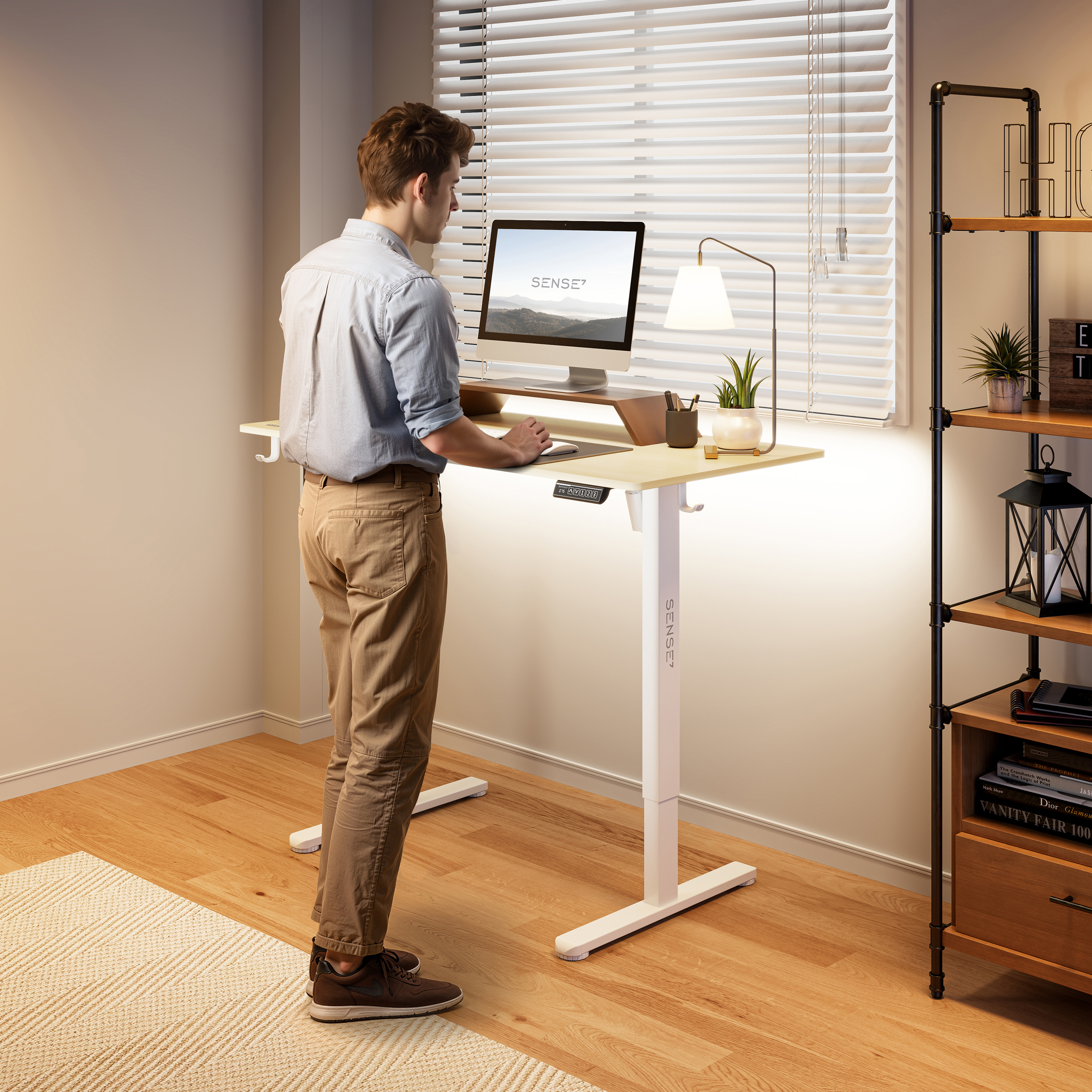 Een man staat aan een staande bureau. Een computer, lamp en plant staan op het bureau. Schappen met boeken en decoratie op de achtergrond.