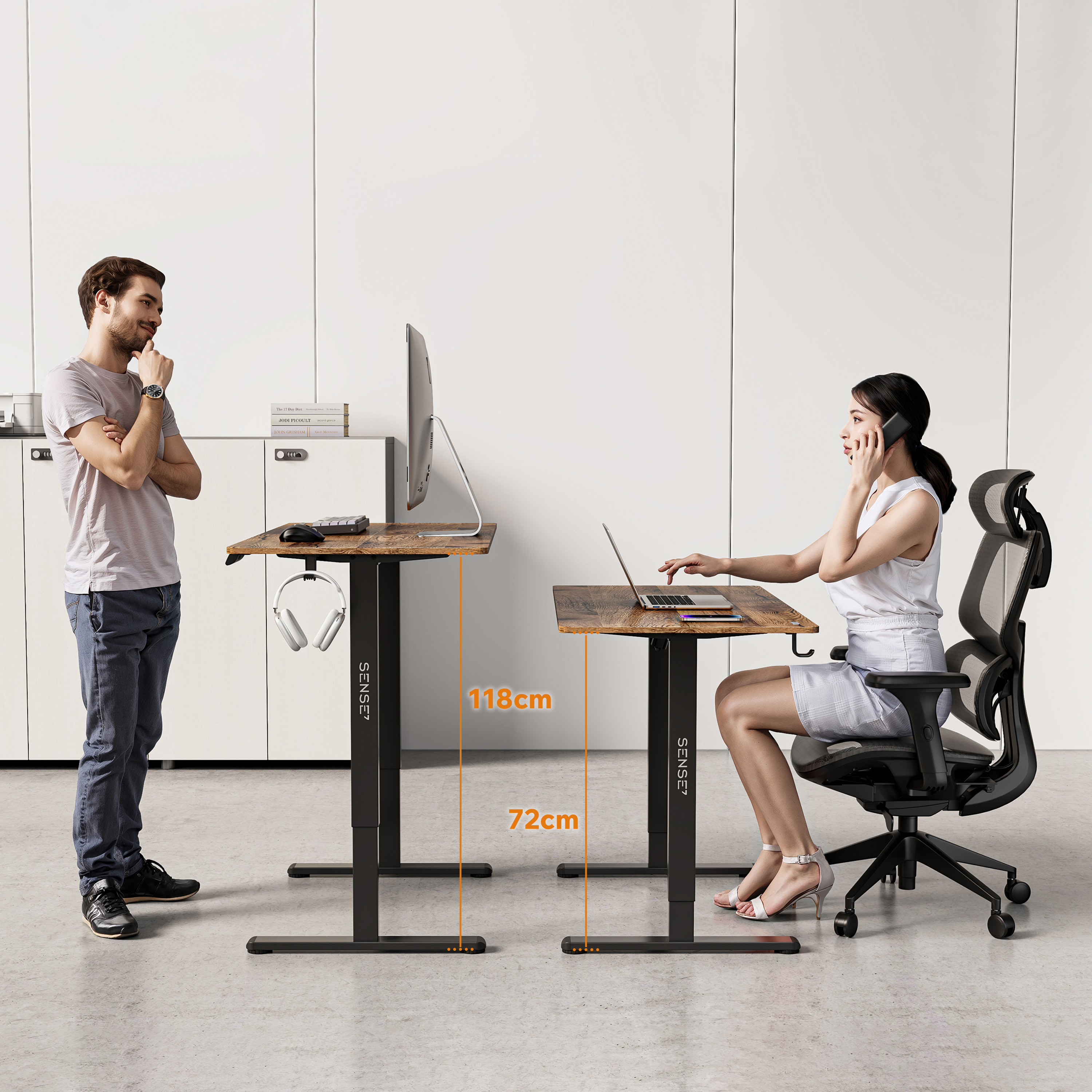 Ein Mann und eine Frau stehen in der Nähe von Schreibtischen. Die Frau telefoniert. Der Mann schaut die Frau an. Beide Schreibtische haben einen Monitor.