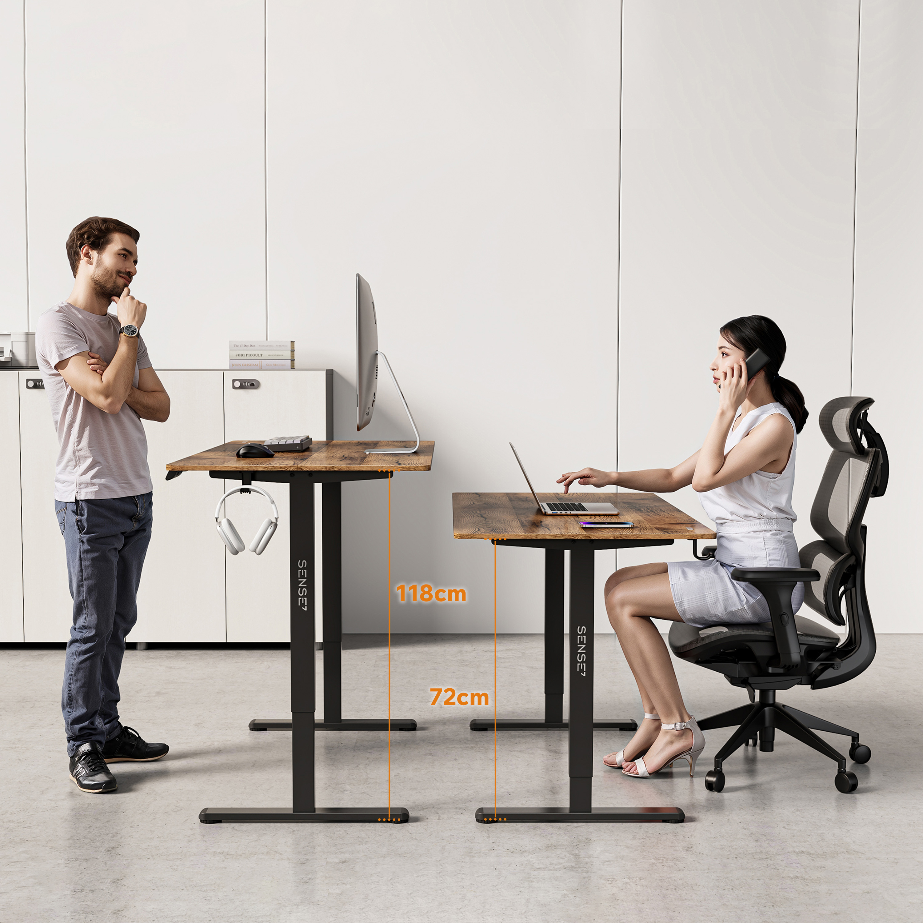 Een man en een vrouw aan bureaus. De ene staat en kijkt naar de andere, die zit en aan het bellen is. Bureaus hebben monitoren.