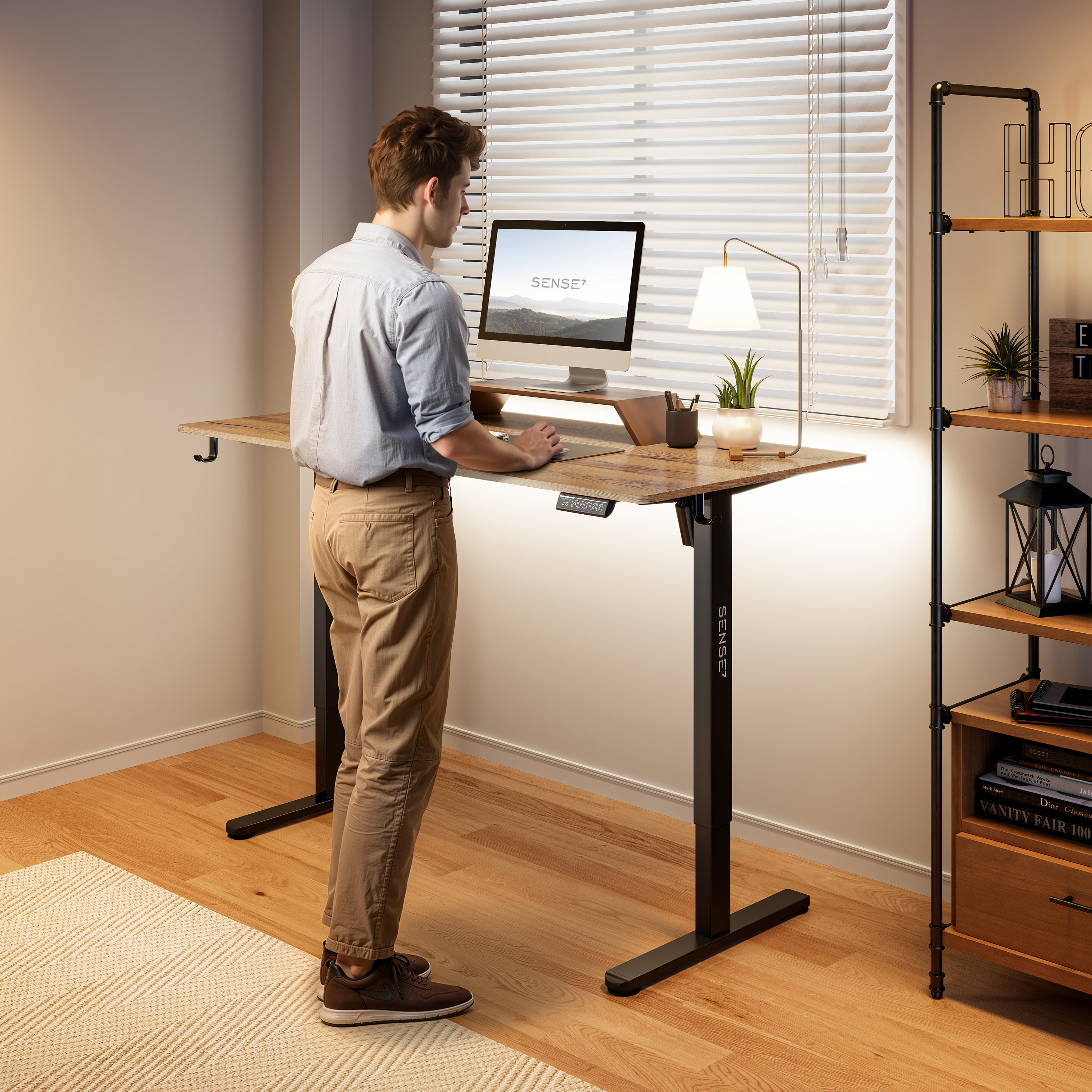 Man staat aan een verstelbaar bureau met een computer. Er is een lamp, een plank en een raam. De vloer is van hout.