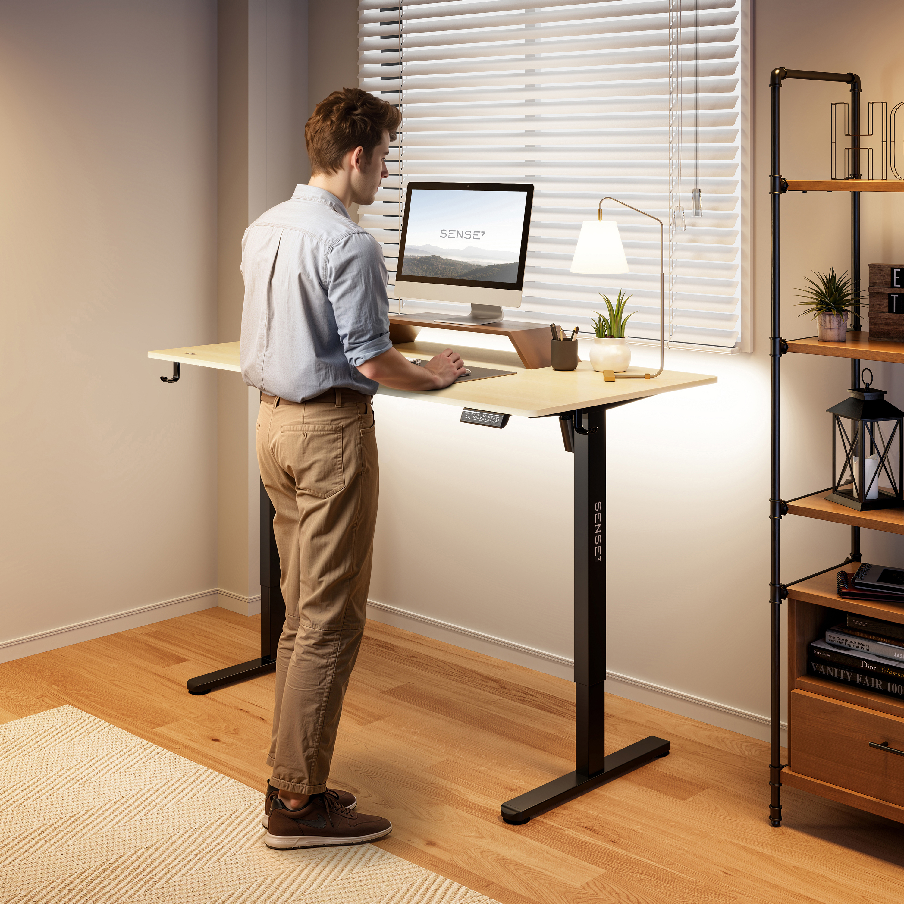 Een man staat aan een verstelbaar bureau met een monitor. Er is een lamp, een plank en een raam met jaloezieën op de achtergrond.